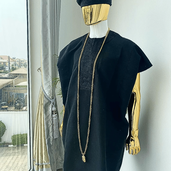 A charcoal black aso dashiki with a floral and box-shaped embroidery design, a hand-weaved Yoruba hat, and a gold Jesus timepiece